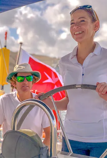 A smiling sailing instructor steering a yacht with a crew member beside her, set against a bright sky and a red maritime flag, capturing a hands-on training moment at sea.
